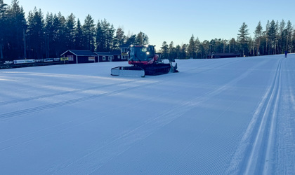 Pistmaskin preparerar ett brett skidspår på snötäckt område med tallskog och byggnader i bakgrunden.