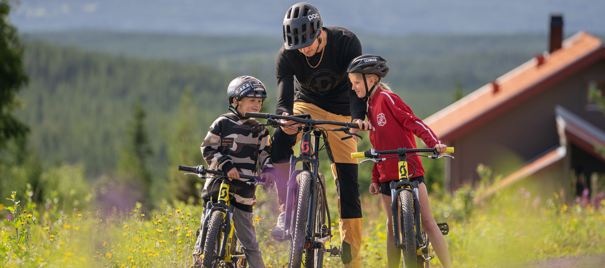 Vuxen och två barn med cyklar på en grusväg i fjällmiljö.