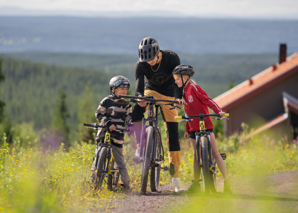 En cykelinstruktör pratar med två barn i hjälm och cykelkläder på en stig med utsikt över granskog och fjärran landskap.