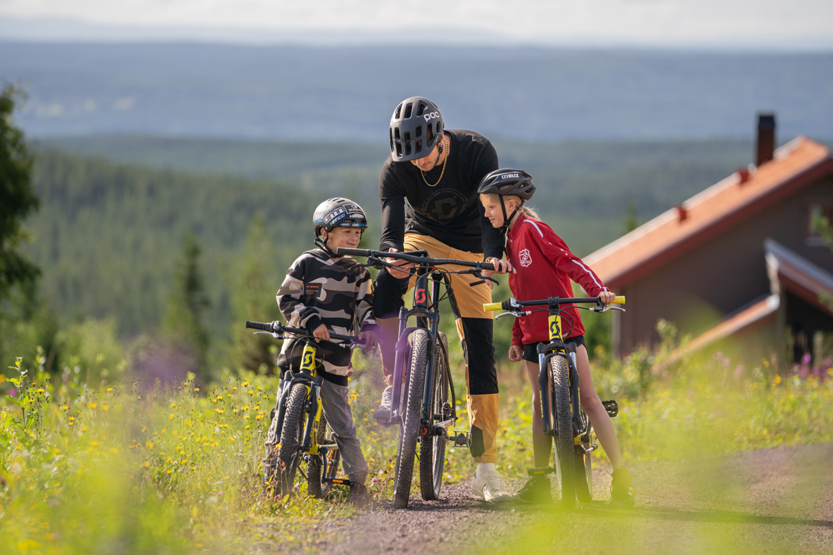 En cykelinstruktör pratar med två barn i hjälm och cykelkläder på en stig med utsikt över granskog och fjärran landskap.