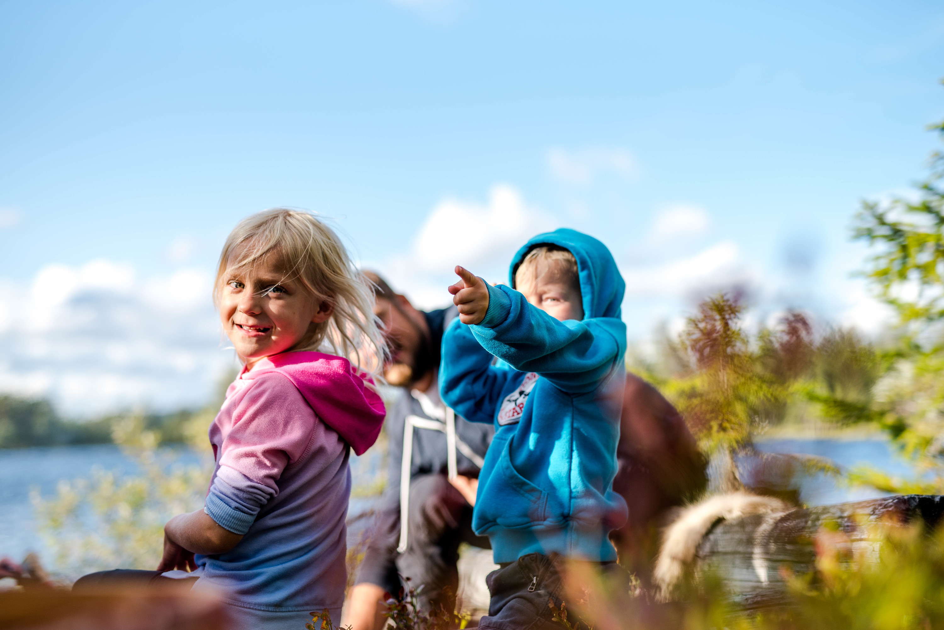 Två barn sitter vid en sjö i solskenet – det ena barnet ler mot kameran medan det andra pekar mot horisonten.