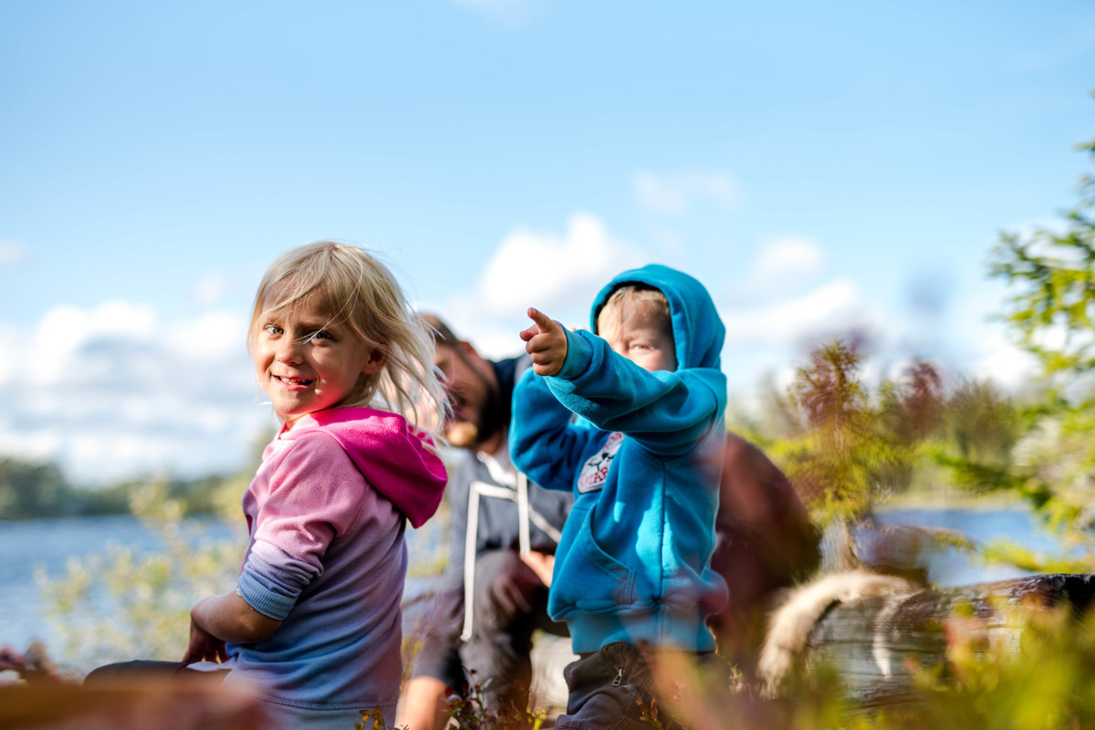 Two children sit by a lake in the sunshine – one smiles at the camera while the other points toward the horizon.
