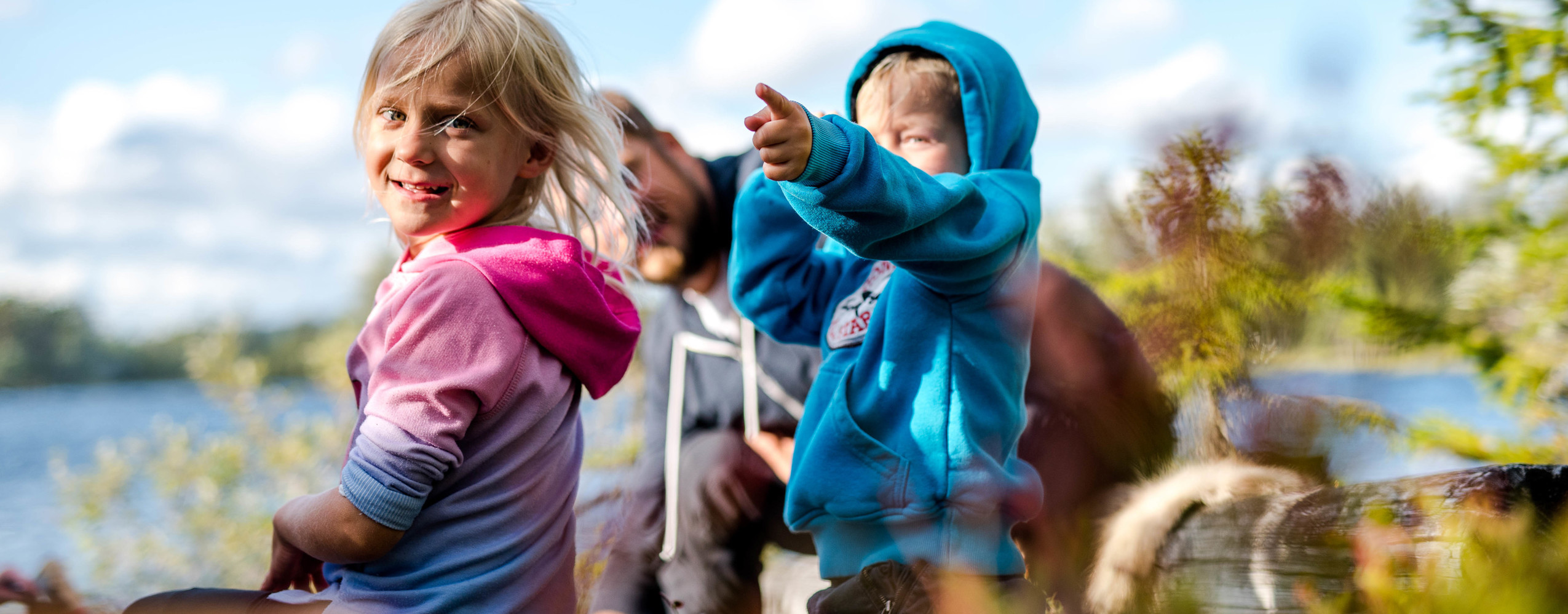 Two children sit by a lake in the sunshine – one smiles at the camera while the other points toward the horizon.