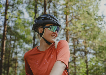En leende kvinna med cykelhjälm och sportglasögon står i skogen med solen som lyser genom träden.