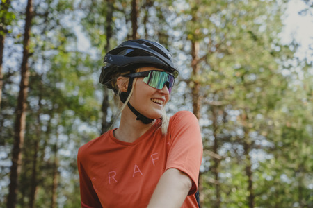 En leende kvinna med cykelhjälm och sportglasögon står i skogen med solen som lyser genom träden.