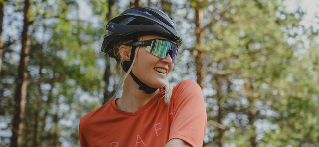 En leende kvinna med cykelhjälm och sportglasögon står i skogen med solen som lyser genom träden.