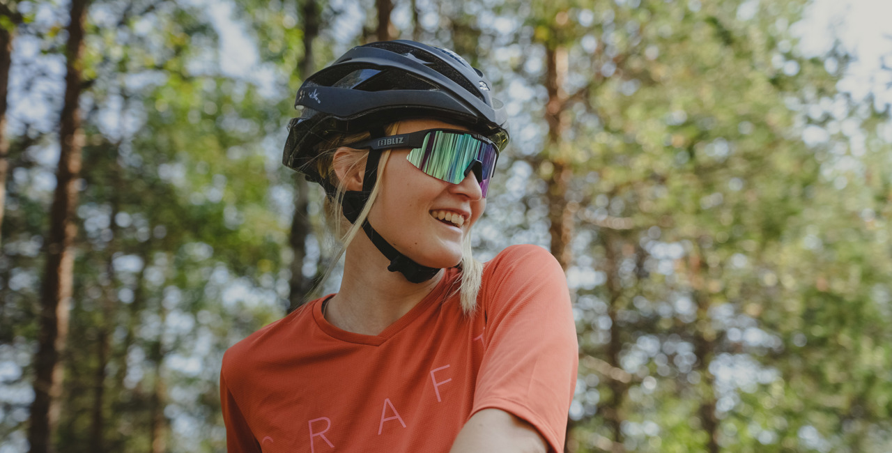 En leende kvinna med cykelhjälm och sportglasögon står i skogen med solen som lyser genom träden.