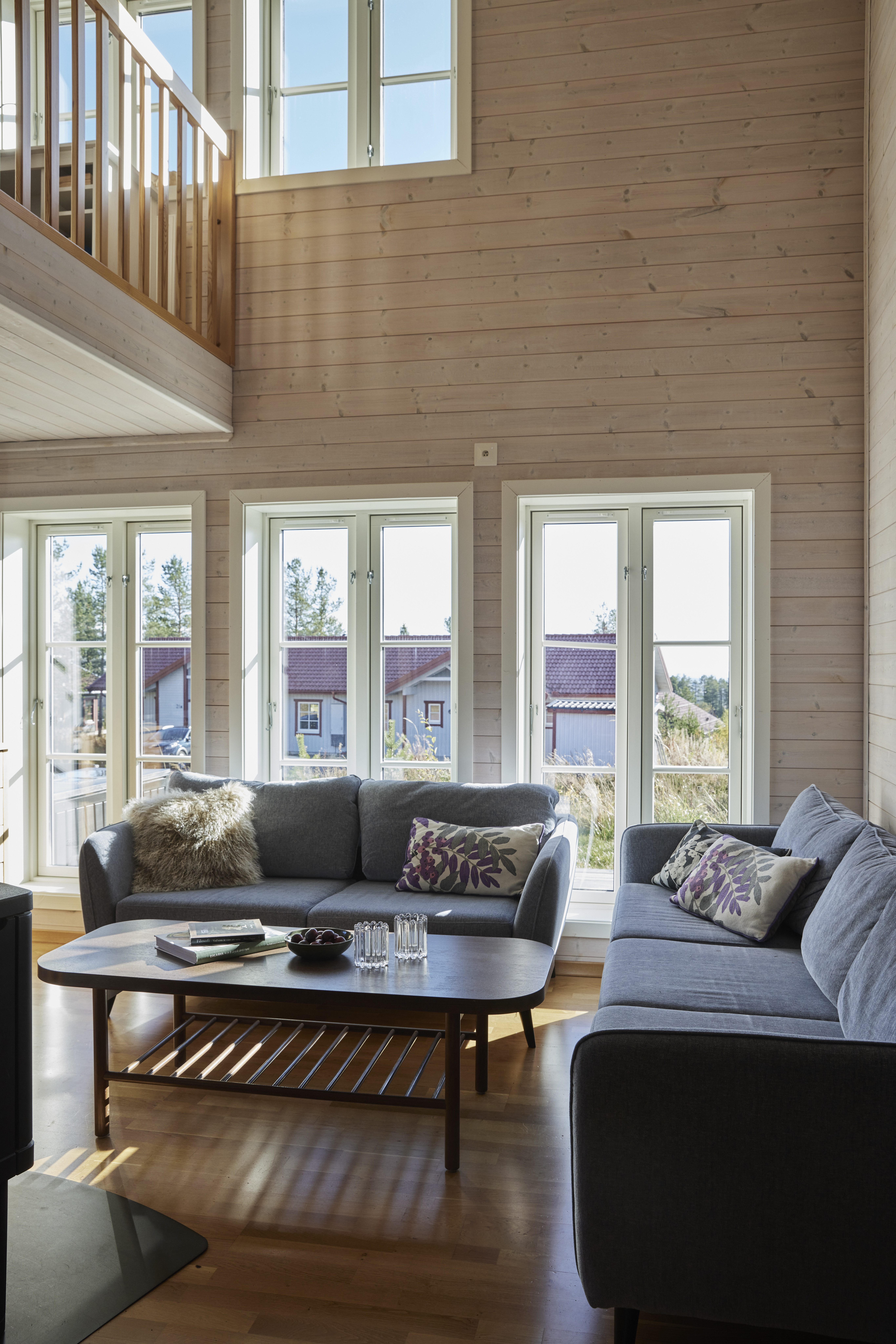 Et lyst opholdsrum med to grå sofaer, et sofabord og store vinduer med udsigt til andre hytter.