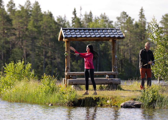 En kvinde kaster sin fiskestang ud ved en rolig sø, mens en mand står ved siden af.