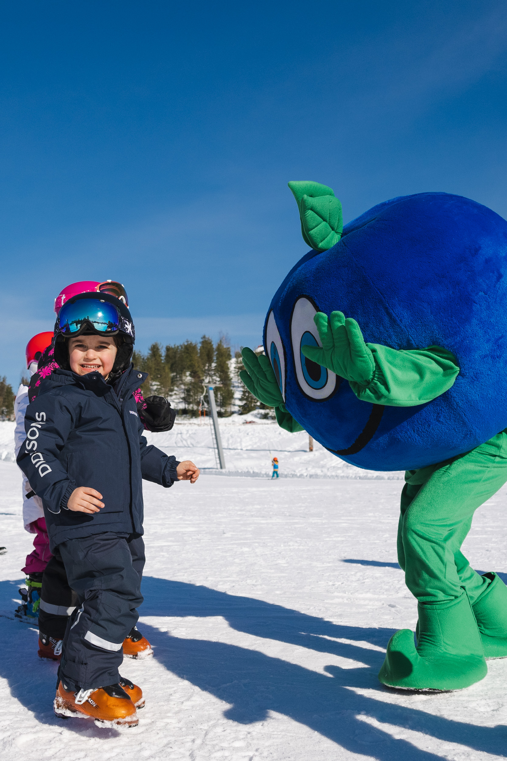 A child in ski gear stands on skis and laughs as the Bärra mascot waves cheerfully on the slope.
