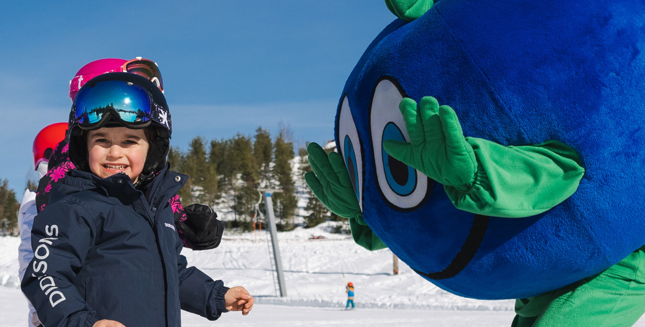 A child in ski gear stands on skis and laughs as the Bärra mascot waves cheerfully on the slope.