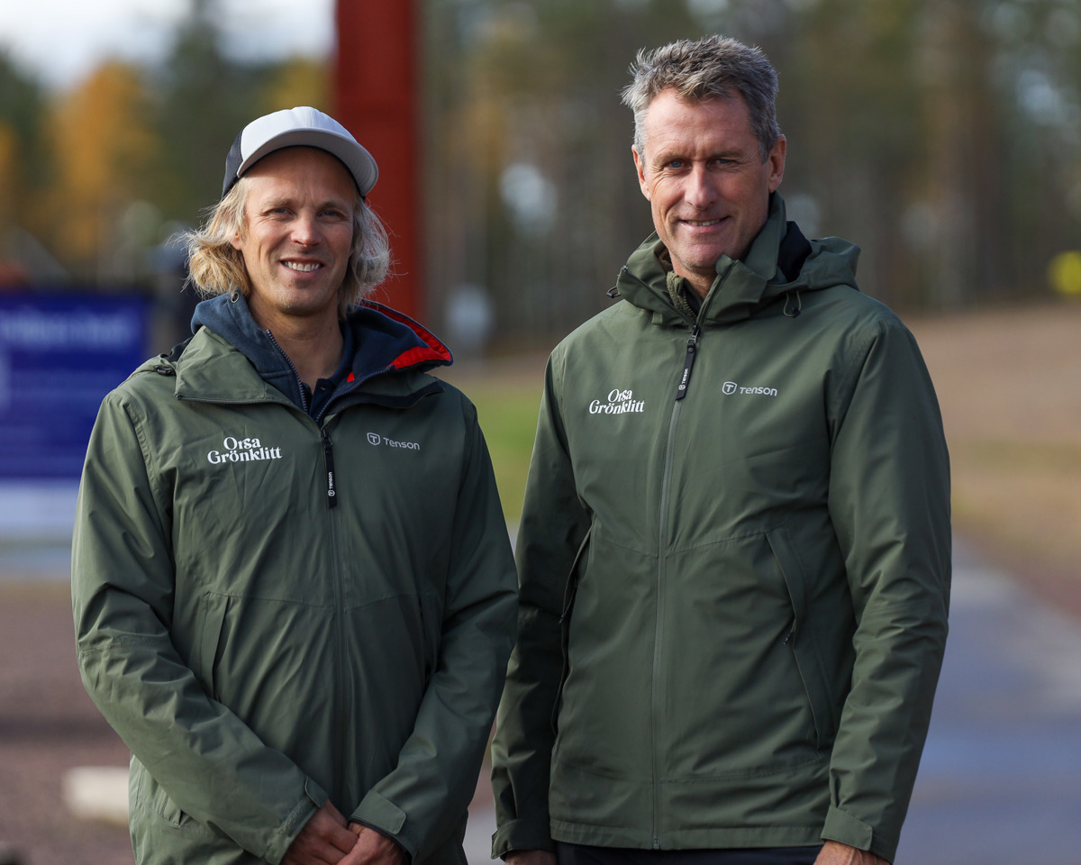 Två personer utomhus i gröna jackor med Orsa Grönklitt-logga, stående på en gångväg med träd i bakgrunden.