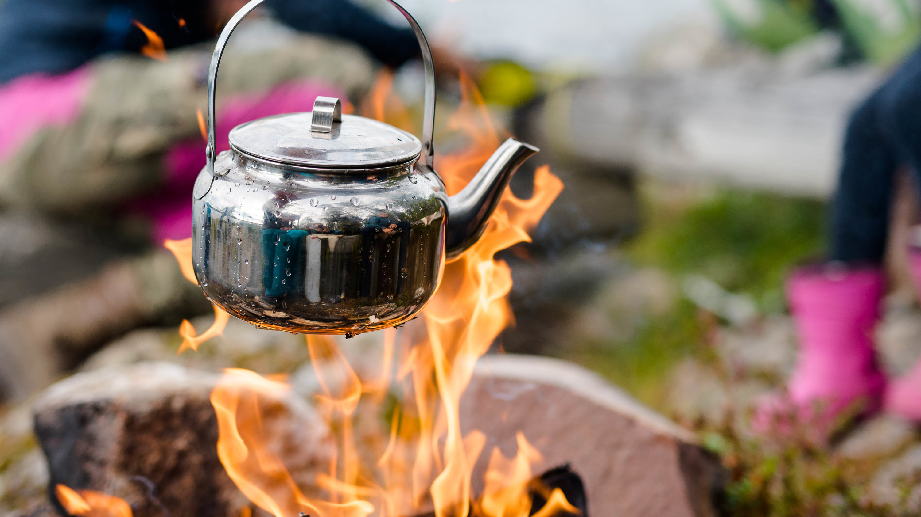Coffee pot hanging over open fire with a woman in colorful clothes in the background.