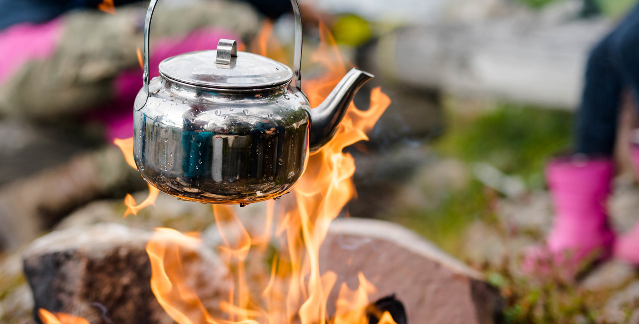 Kaffepanna hängande över öppen eld med kvinna i färgglada kläder i bakgrunden.