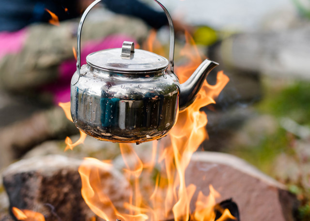 Coffee pot hanging over open fire with a woman in colorful clothes in the background.