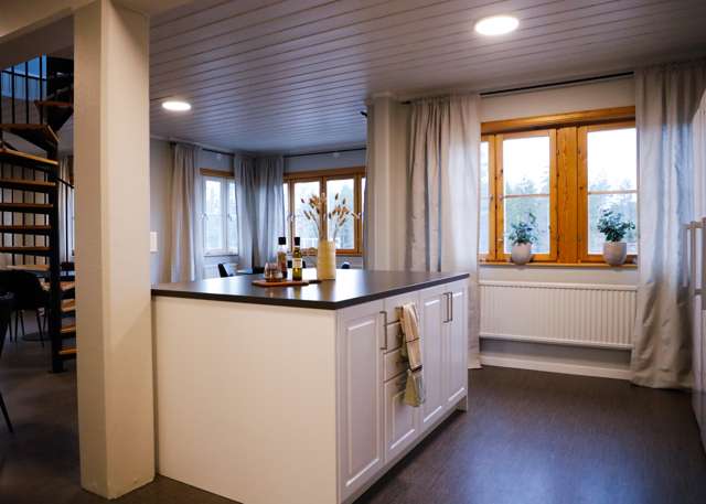 Lyst køkkenområde med køkkenø, vinduer med gardiner og vindeltrappe i baggrunden.