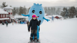 Barn på alpina skidor tillsammans med Bärra Blåbär i ett snötäckt vinterlandskap.