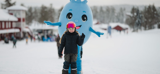Barn på alpina skidor tillsammans med Bärra Blåbär i ett snötäckt vinterlandskap.