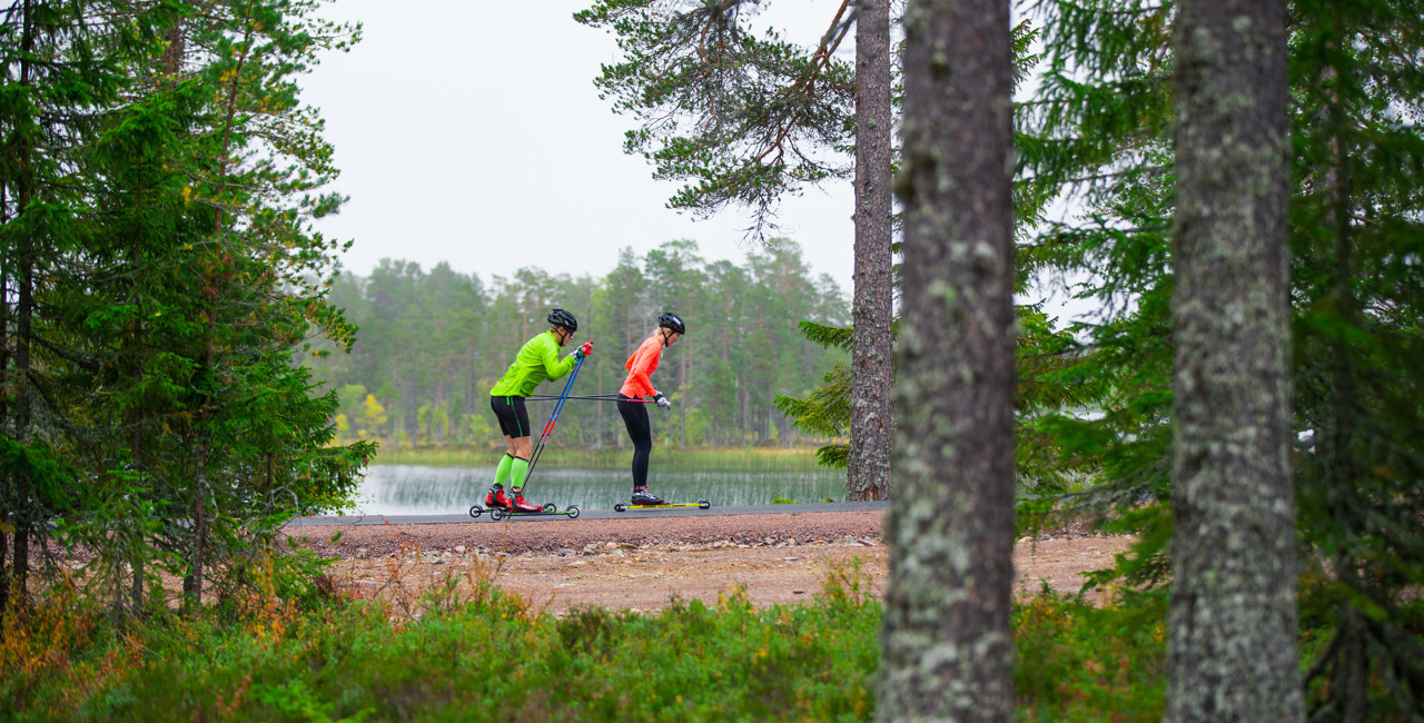 Två personer åker rullskidor på en asfalterad multibana vid en sjö, omgivna av tät granskog.