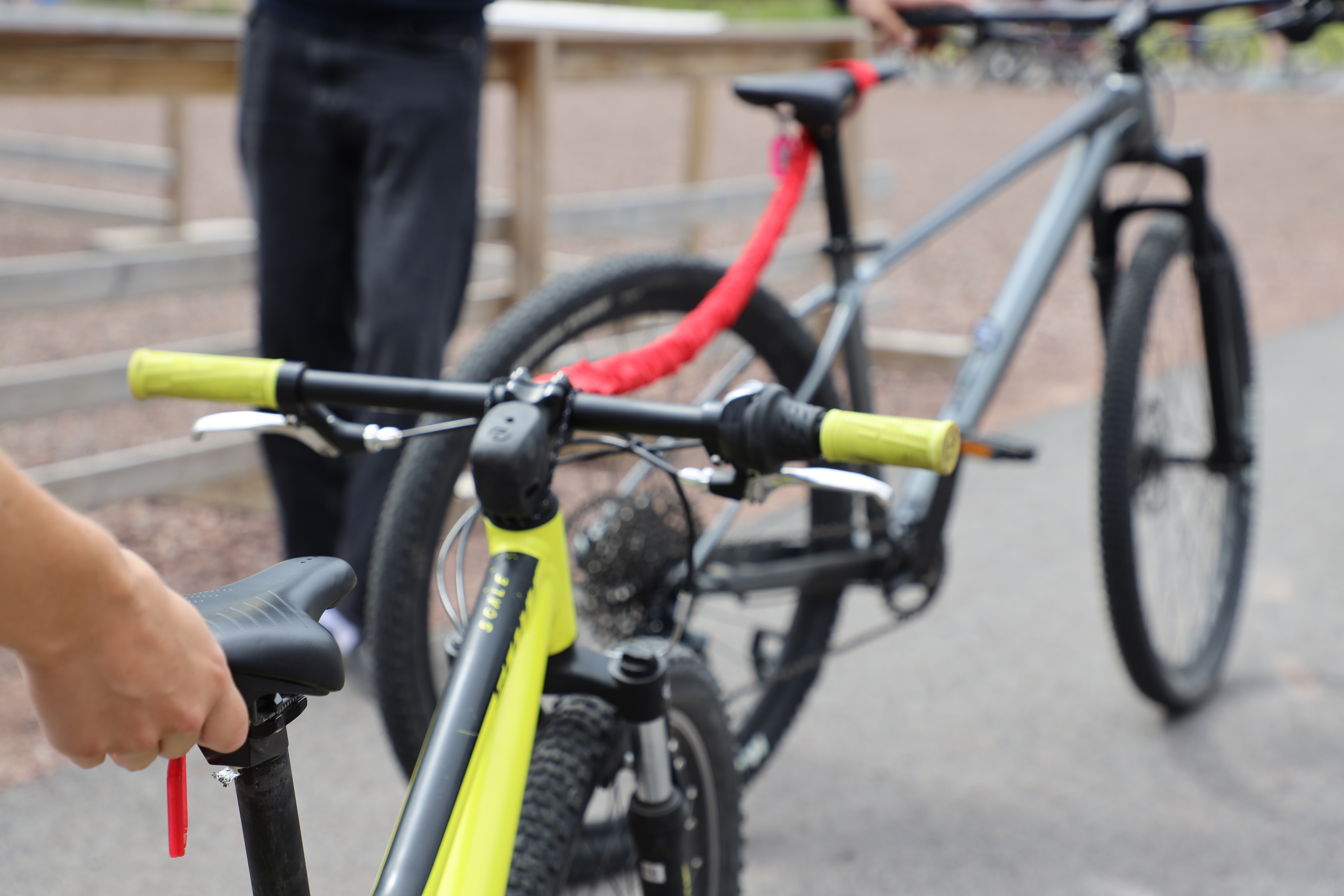 Två mountainbikes kopplade ihop med ett rött bogserband; den främre cykeln är gul och den bakre grå, med personer i bakgrunden på en grusplan.