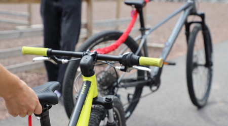 Två mountainbikes kopplade ihop med ett rött bogserband; den främre cykeln är gul och den bakre grå, med personer i bakgrunden på en grusplan.