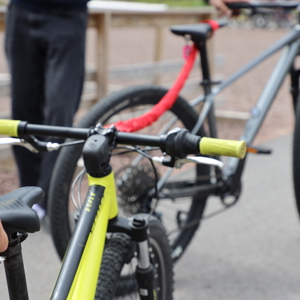 Two mountain bikes connected by a red tow strap; the front bike is yellow and the rear one gray, with people in the background on a gravel area.