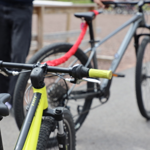 Två mountainbikes kopplade ihop med ett rött bogserband; den främre cykeln är gul och den bakre grå, med personer i bakgrunden på en grusplan.