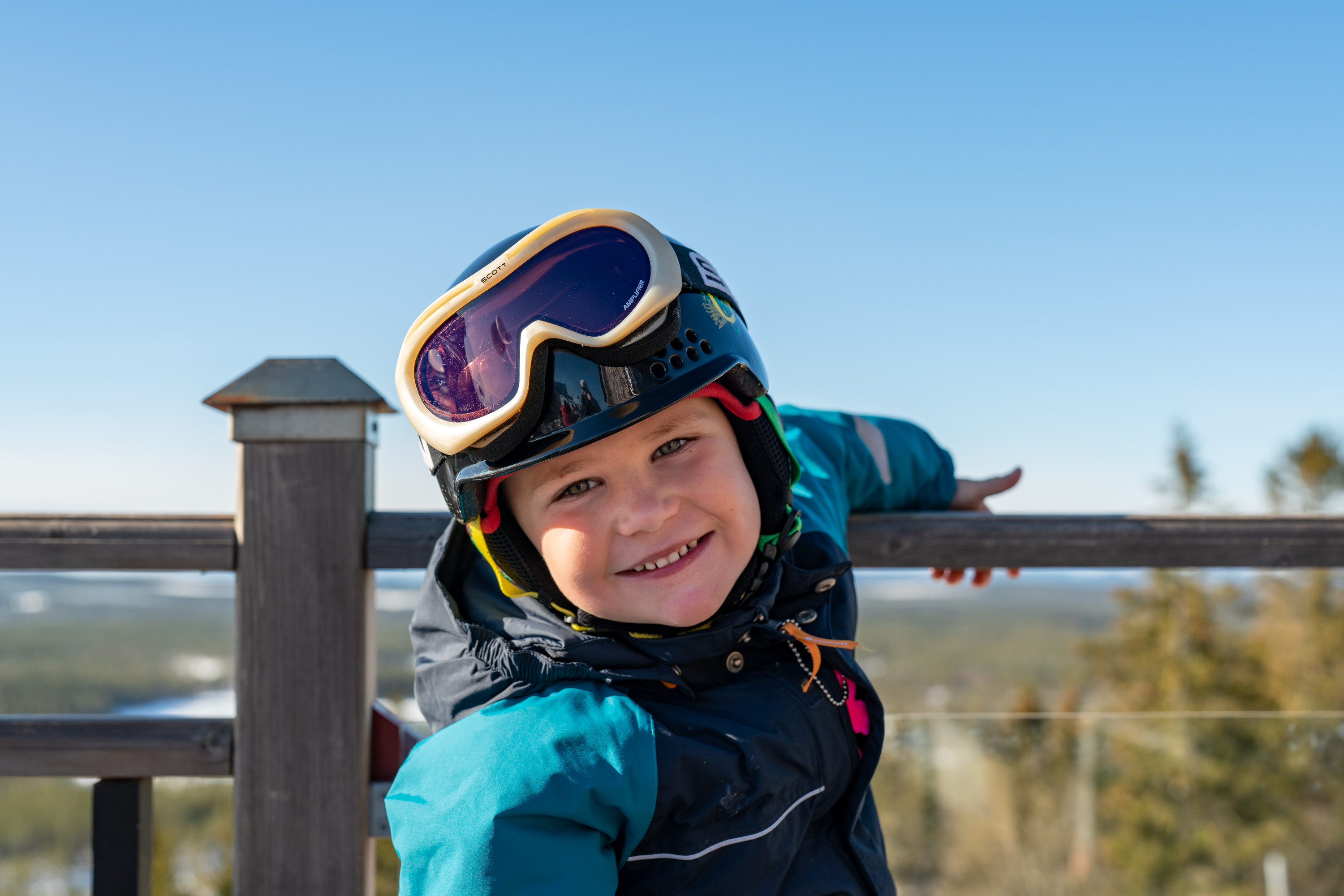 Ett barn i blå skidjacka och hjälm med skidglasögon ler mot kameran vid ett räcke med utsikt över skog och fjäll.