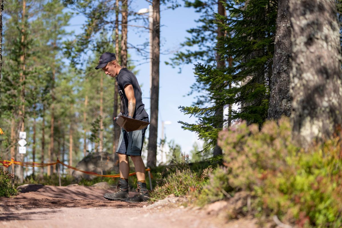 A man is maintaining a bike trail in the forest. He holds a shovel and shapes the ground in a sunlit pine forest.