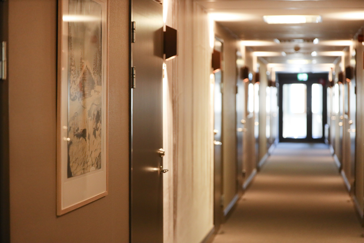 Lång korridor med stängda dörrar, mattgolv och varm belysning.