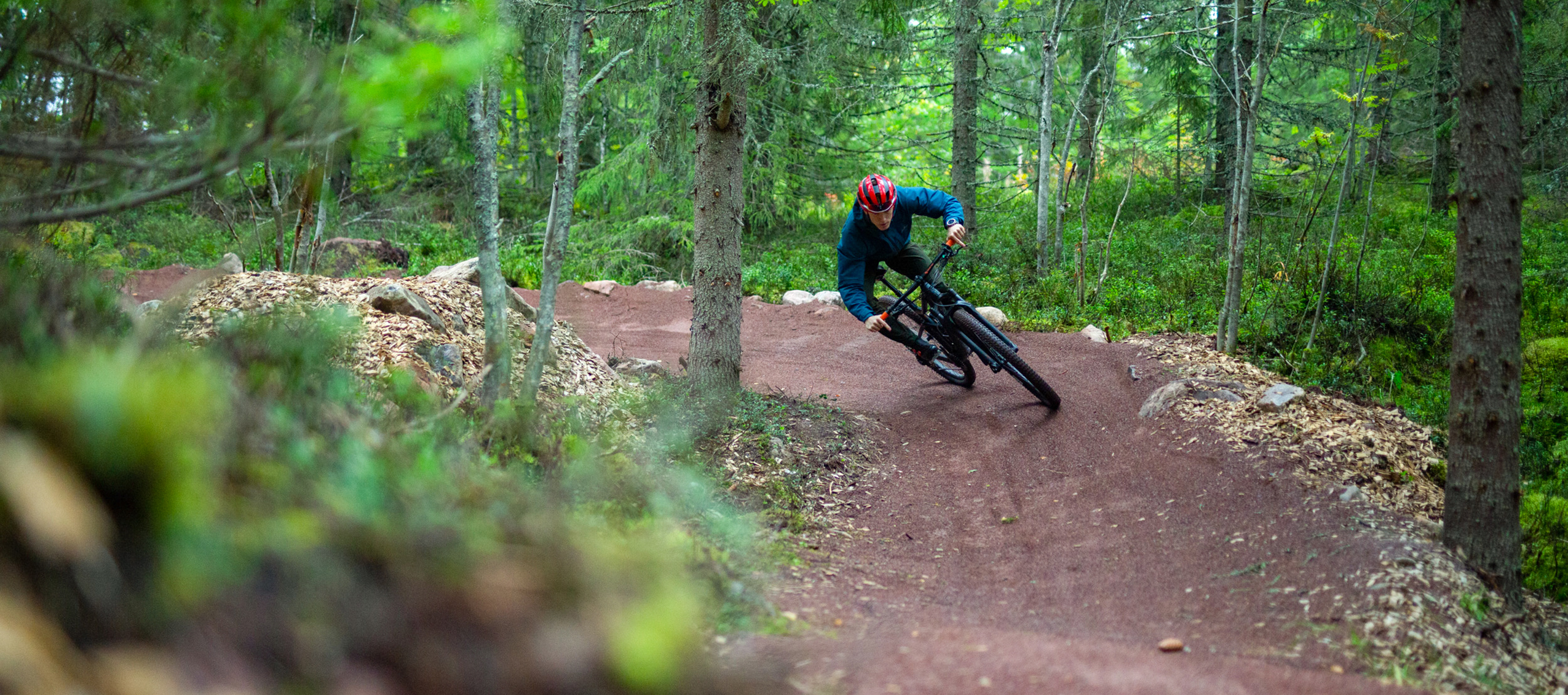 Cyklist med hjälm susar fram i en kurva på en röd grusad mountainbikeled i en grön och tät skog.