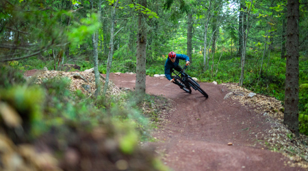En cyklist med hjelm suser gennem en kurve på en rød grusbelagt mountainbikerute i en tæt, grøn skov.