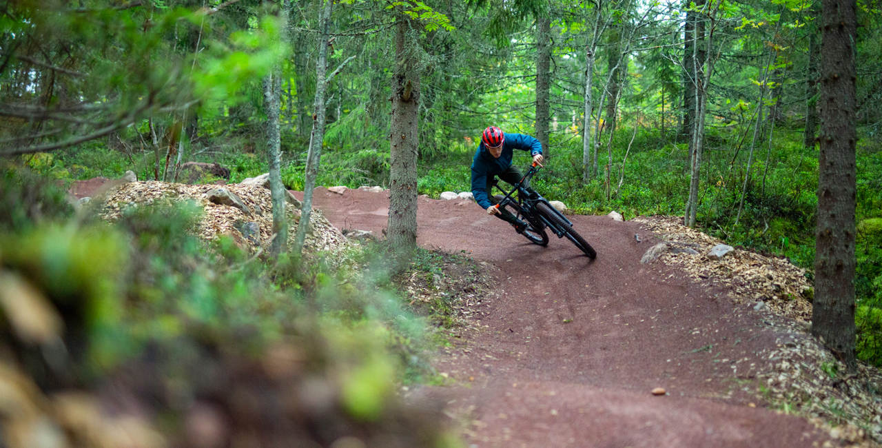 A cyclist wearing a helmet rides through a curve on a red gravel mountain bike trail in a dense green forest.