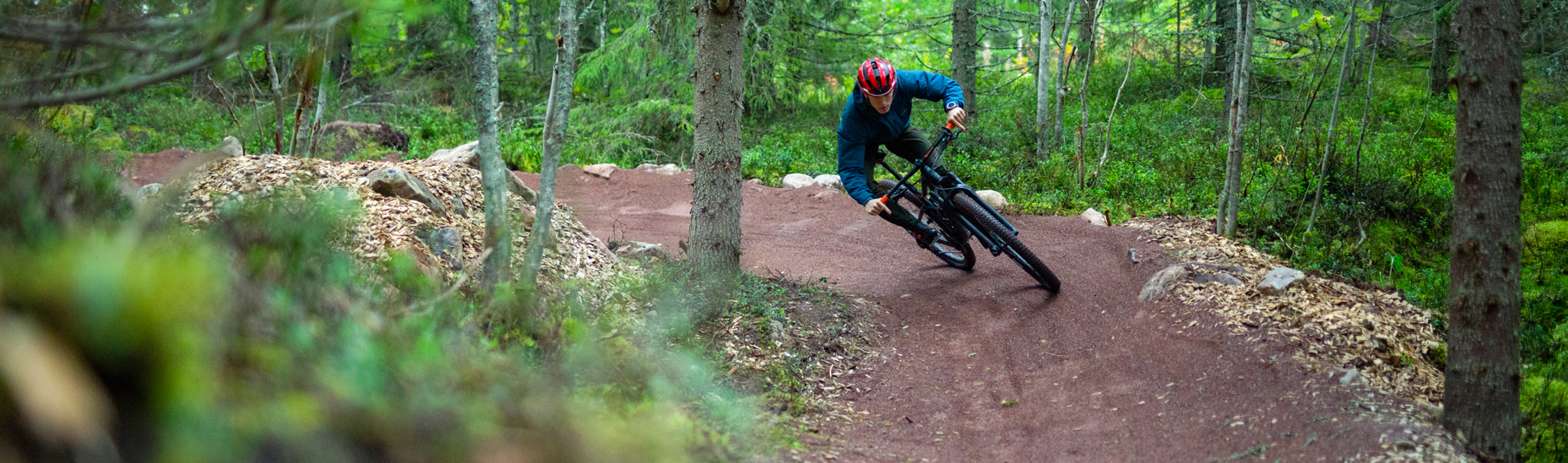 A cyclist wearing a helmet rides through a curve on a red gravel mountain bike trail in a dense green forest.