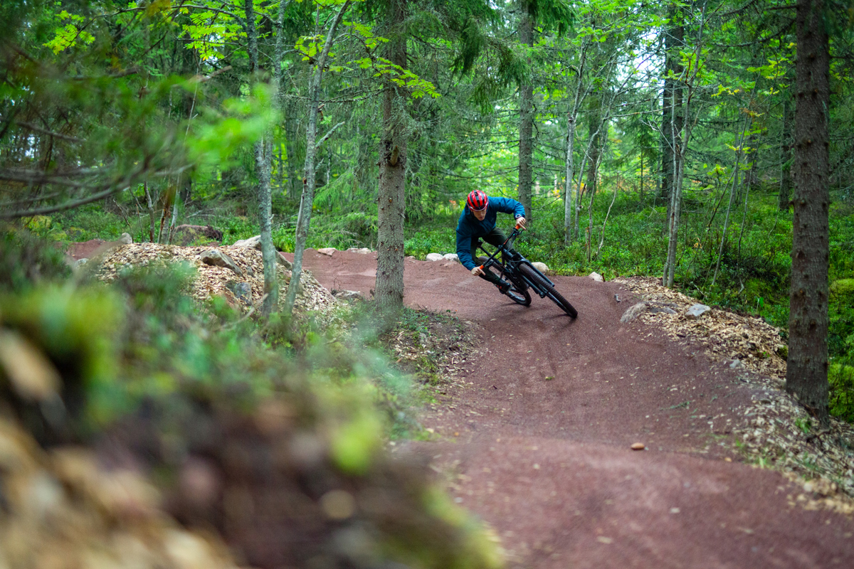En cyklist med hjelm suser gennem en kurve på en rød grusbelagt mountainbikerute i en tæt, grøn skov.