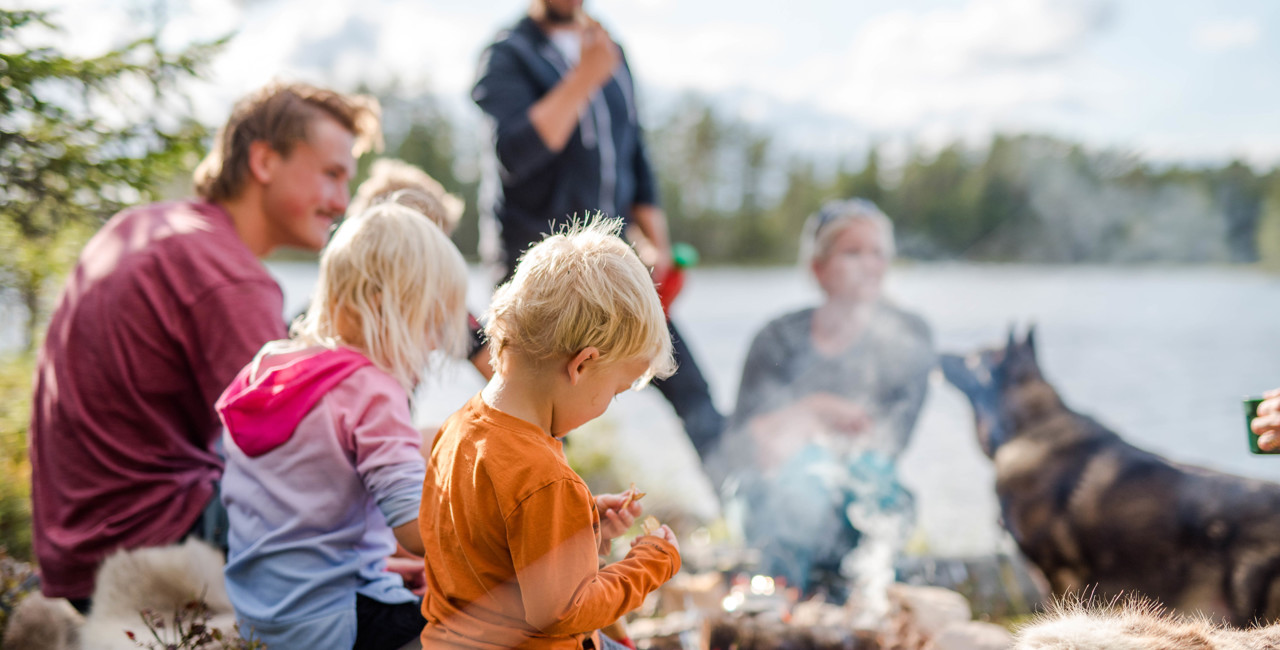 Familj med barn som grillar vid en öppen eld i naturen nära en sjö, med en hund i bakgrunden.
