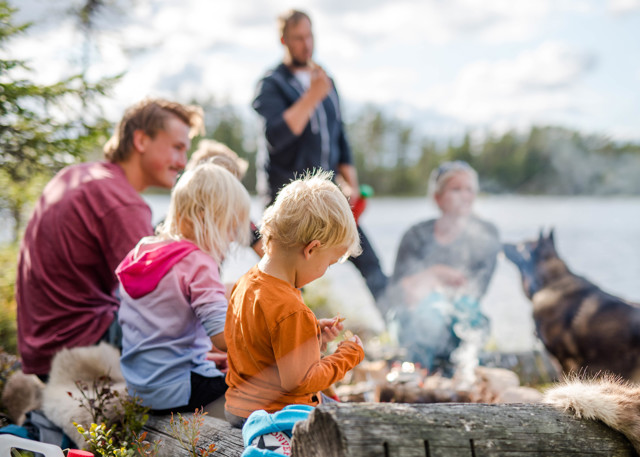 Family with children grilling by an open fire in nature near a lake, with a dog in the background.