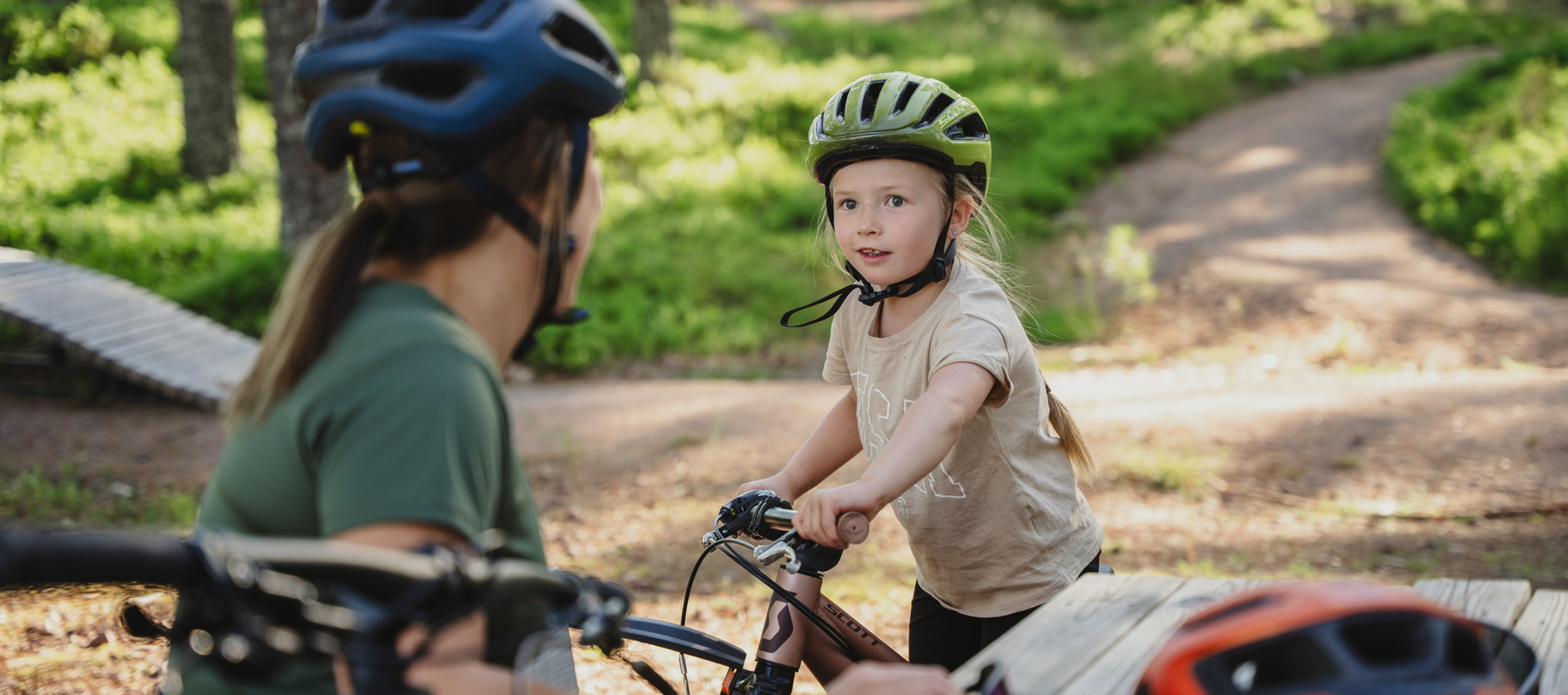 Ett barn med cykelhjälm står med sin cykel och pratar med en vuxen vid en bänk i en skogsmiljö med cykelstigar.