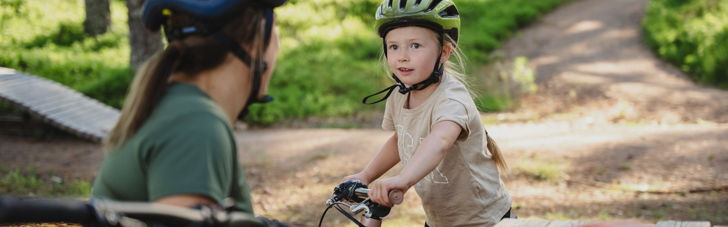 A child wearing a bike helmet stands with their bicycle and talks to an adult by a bench in a forest setting with bike trails.