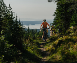Personer cyklar mountainbike längs en smal skogsstig med utsikt över en sjö i bakgrunden.
