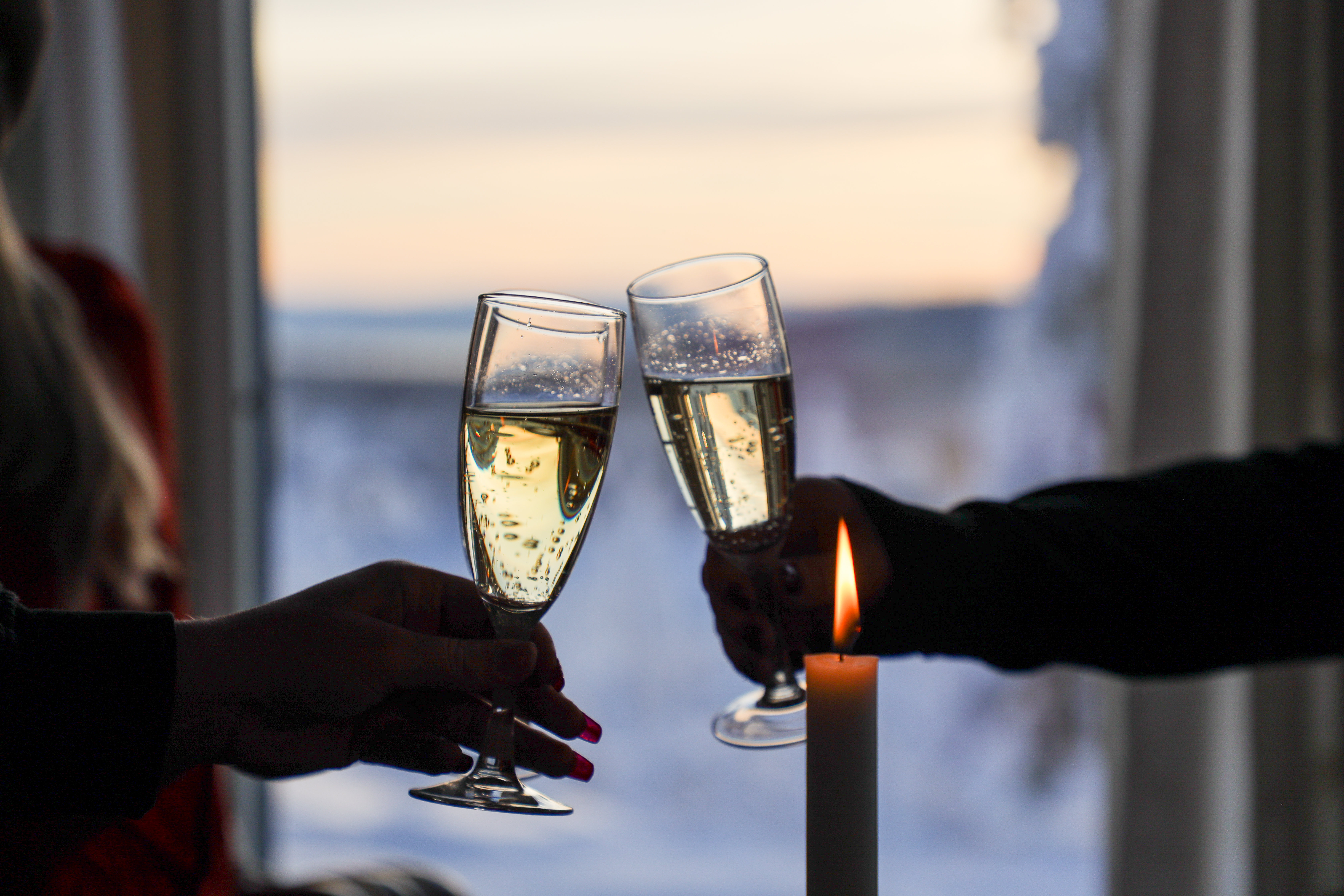 Två personer skålar med champagneglas vid ett fönster med vinterutsikt och levande ljus.