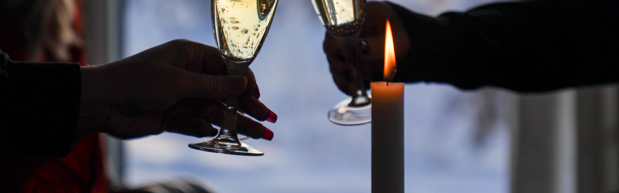 Two people clinking champagne glasses by a window with a winter view and a lit candle.