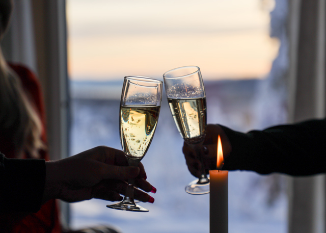Två personer skålar med champagneglas vid ett fönster med vinterutsikt och levande ljus.
