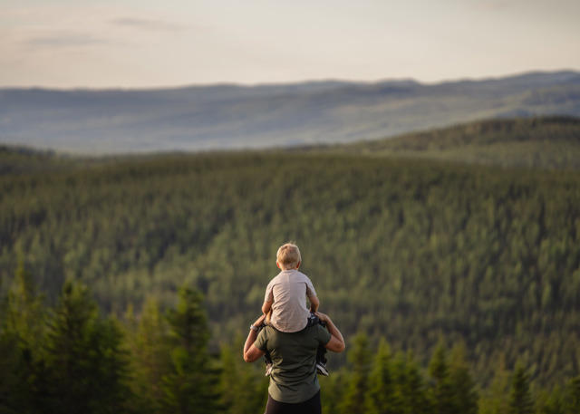 Vuxen bär barn på axlarna med utsikt över skogsklädda berg i solnedgång.