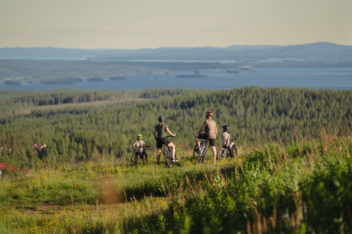 Familj på cyklar blickar ut över skogslandskap och sjöar i kvällsljus.