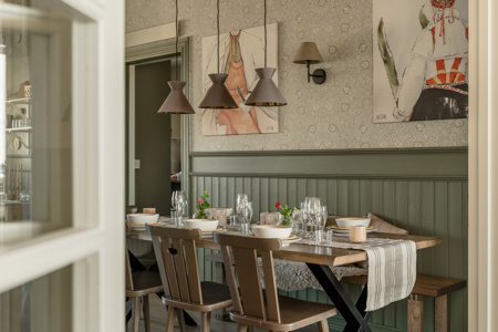 En dukad matplats med träbord, stolar, glas och porslin i ett rum med målningar och hängande lampor.