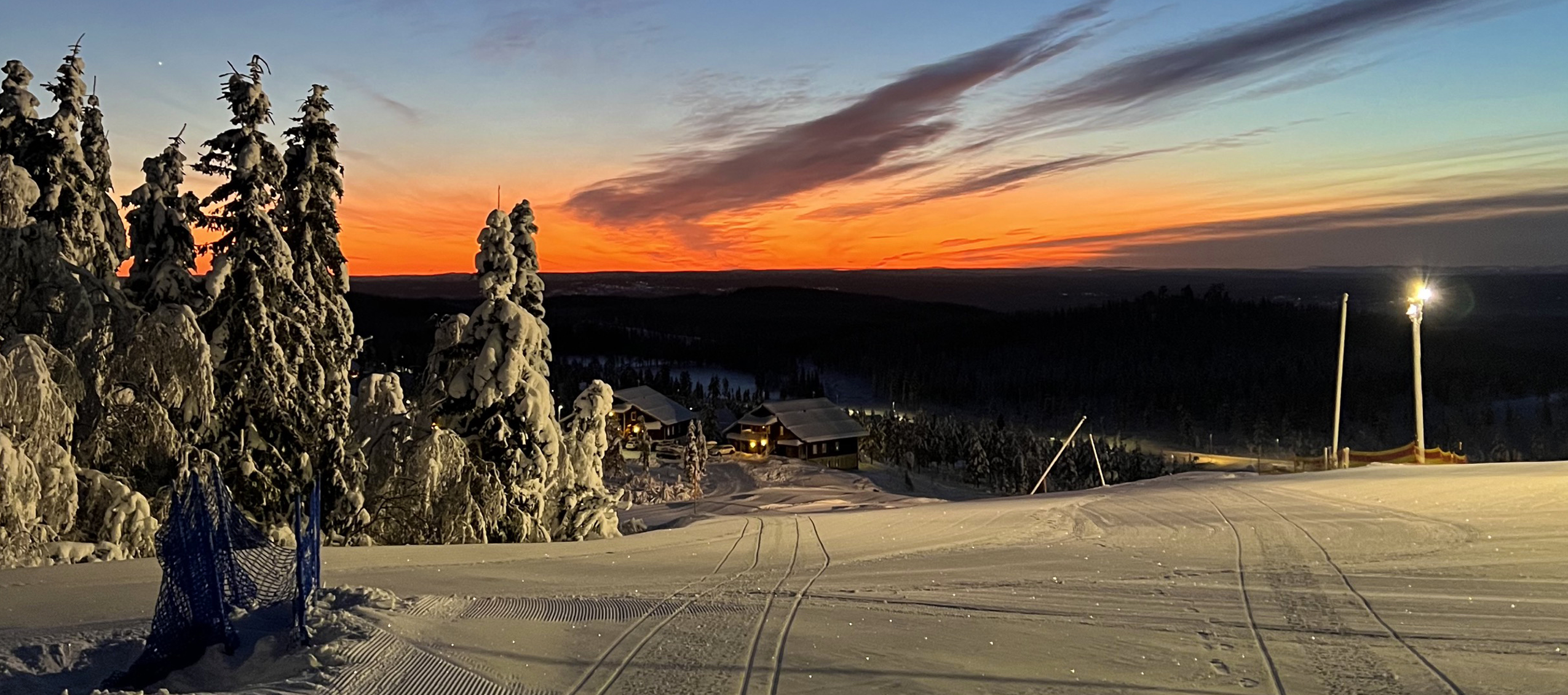 Snötäckt skidbacke i kvällsljus med solnedgång och belysta träd.
