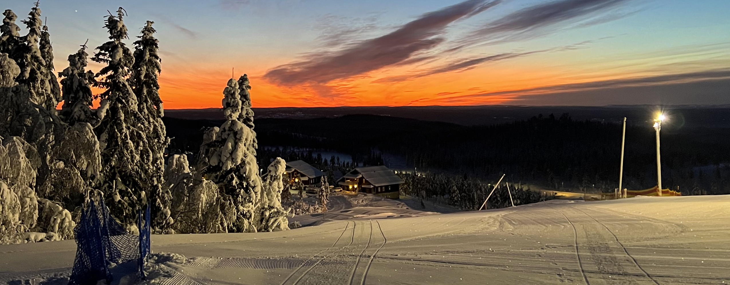 Snow-covered ski slope in evening light with sunset and illuminated trees.