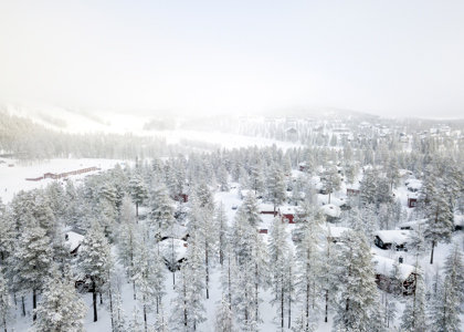 Snedækket hytteområde omgivet af tæt skov i Orsa Grönklitt, med pister og anlæg i baggrunden i tåget vinterlys.