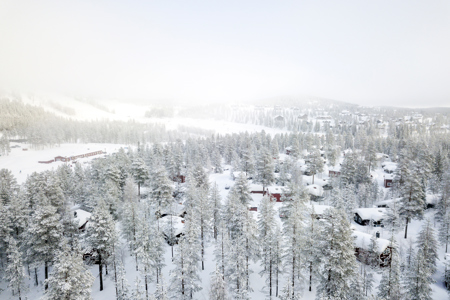 Snötäckt stugområde omgivet av tät skog i Orsa Grönklitt, med backar och anläggning i bakgrunden i dimmigt vinterljus.